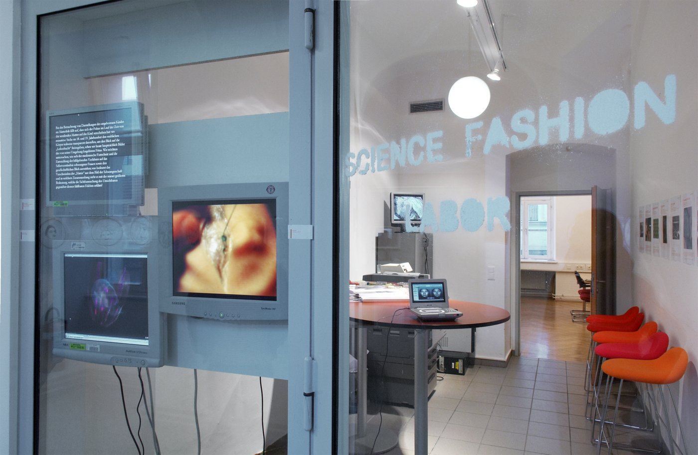 Man sieht den Eingang zum Science Fashion Labor mit mehreren Monitoren, einem Tisch und orangen Sesseln.