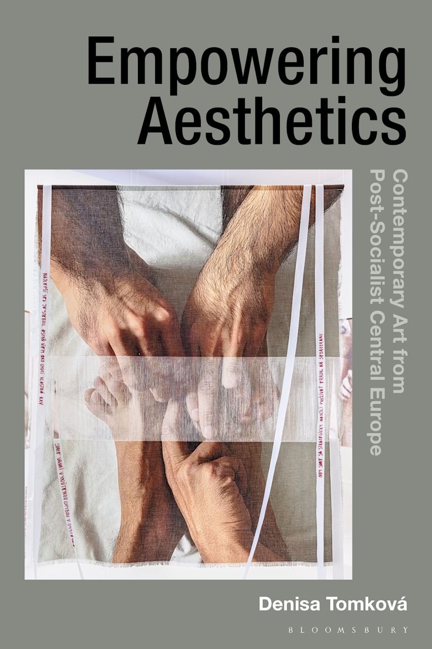 Book cover of "Empowering Aesthetics: Contemporary Art from Post-Socialist Central Europe" by Denisa Tomková. Against a gray background, the title is in large black text at the top, with the author and Bloomsbury logo at the bottom. The subtitle runs vertically in white text on the right. The central artwork shows intertwined hands and feet printed on a creased, translucent strip. This strip, which has fragmented text along its edges, rests on a light, textured surface.