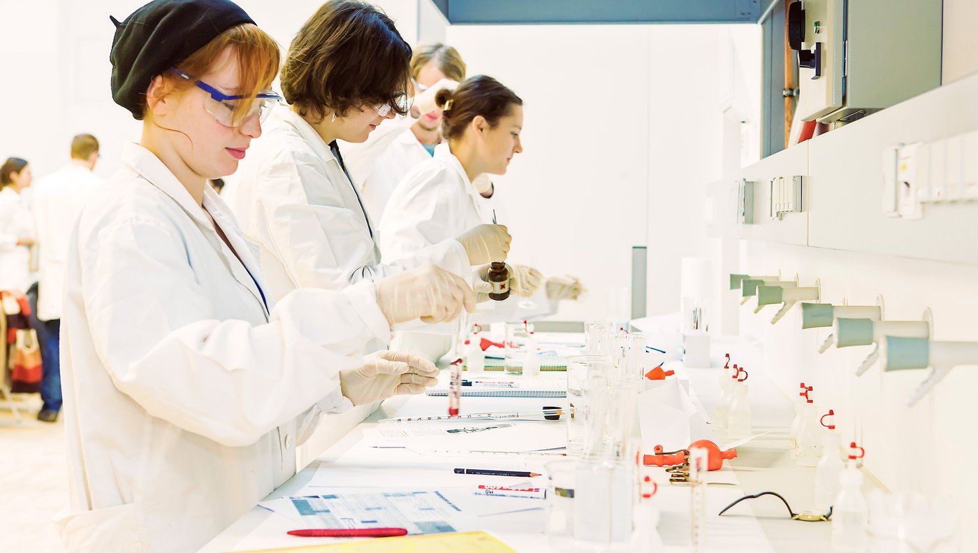 Studierende in weißen Laborkitteln stehen an einer Laborbank.Eine Personhält ein Probenröhrchen, eine andere ein braunes Fläschen in den Händen.