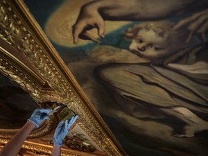 Nahaufnahme des Deckengemäldes der Aula, eine Hand mit Schwamm reinigt den Goldrahmen um das Gemälde