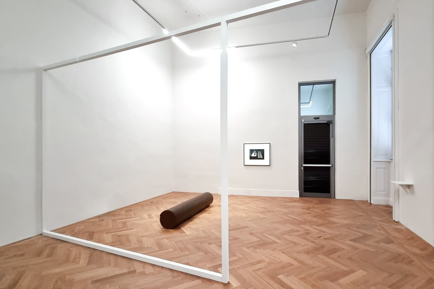 Ausstellungsansicht eines Raumes mit einem weißen Gestell, eine braune Holzrolle am Boden, im Hintergrund ein schwarz-weißes Bild an der Wand und eine geschlossene Türe