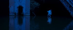 Ein weiß geschminkter und gekleidedeter Mann in einer Geste des Butoh-Tanzes steht in einem See innerhalb eines verfallenen Gebäudes.