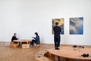 Ausstellungsansicht Raum 3 Personen Tisch und zwei Bilder an der Wand