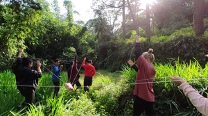 Mehrere Personen in einer grünen Naturlandshcaft, die in einem Kreis gehen, in ihrer Mitte ein Gegenstand, der an Fäden hängt, die die Personen in ihren Händen tragen.