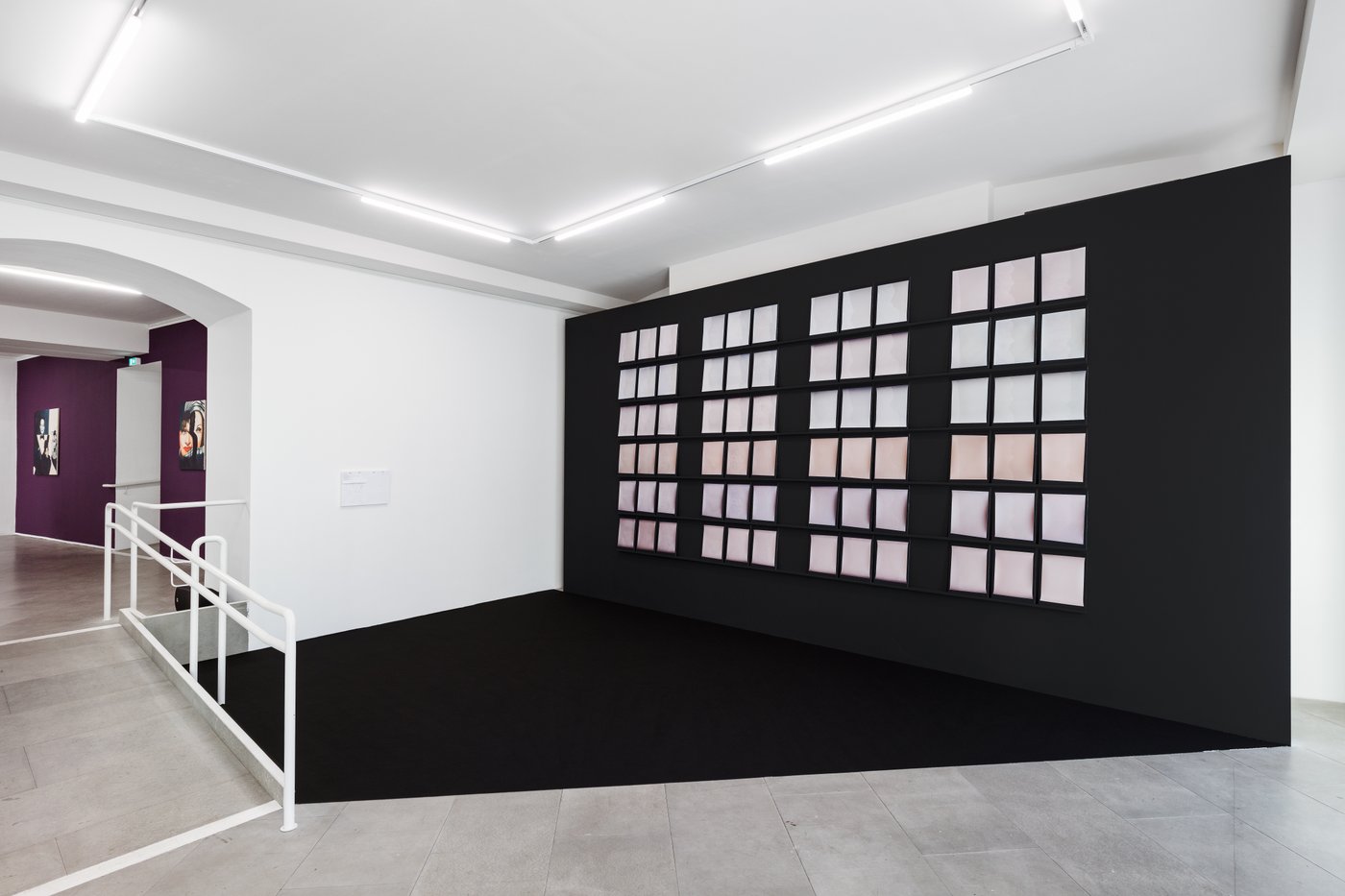 Auf einer Art schwarzen Bühne sieht man 4 Blöcke von jeweils 3 x 6 Reihen von Montioren, die wie rosa Kissen wirken. Im Hintergrund Bilder auf einer lila Wand.