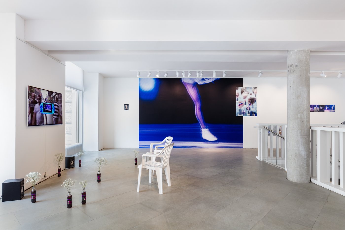 Im Ausstellungsraum stehen vor einem Screen Colaflaschen mit weißen Blüten und zwei Plastikstühle. Im Hintergrund eine Fototapete mit dem Bein einer Schlittschuhläuferin und andere fotografische Auseinandersetzungen mit dem Thema.