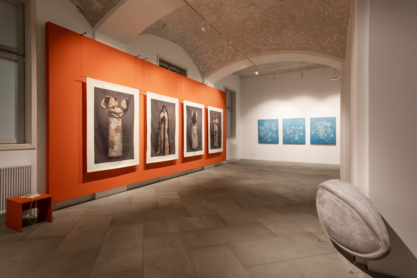 Im Gewölberaum hängen auf der linken Seite vor einer orangen Wand vier Stoffbahnen mit der Abbildung einer Frau in archaisch wirkenden Kleidern. Auf der Rückwand hängen drei blaue abstrakte Malereien, rechts vorne eine linsenförmige Steinskulptur.