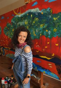 Frau mit dunkelbraunen Locken in einer Jeans-Latzhose und einem blau-weiß gestreiften Shirt, neben ihr eine Kiste mit Malerfarben und im Hintergrund ein großes Gemälde mit rotem Hintergrund und gelben kleinen Fischen und einer grün-blauen wolkenartigen Struktur in die gelbe Punkte und kleine gelbe Vögel gemalt sind