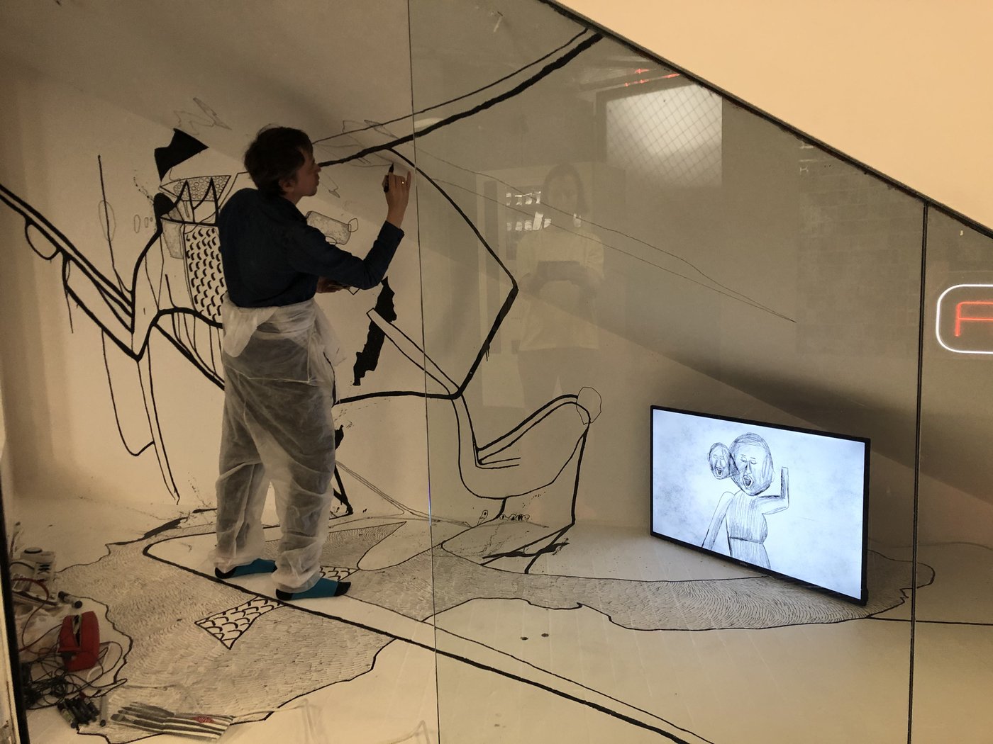 Installationsansicht mit Künstler der gerade dabei ist die Wand mit schwarzem Stift zu bemalen, in der rechten Ecke steht ein Bildschirm mit gezeichneter Figur