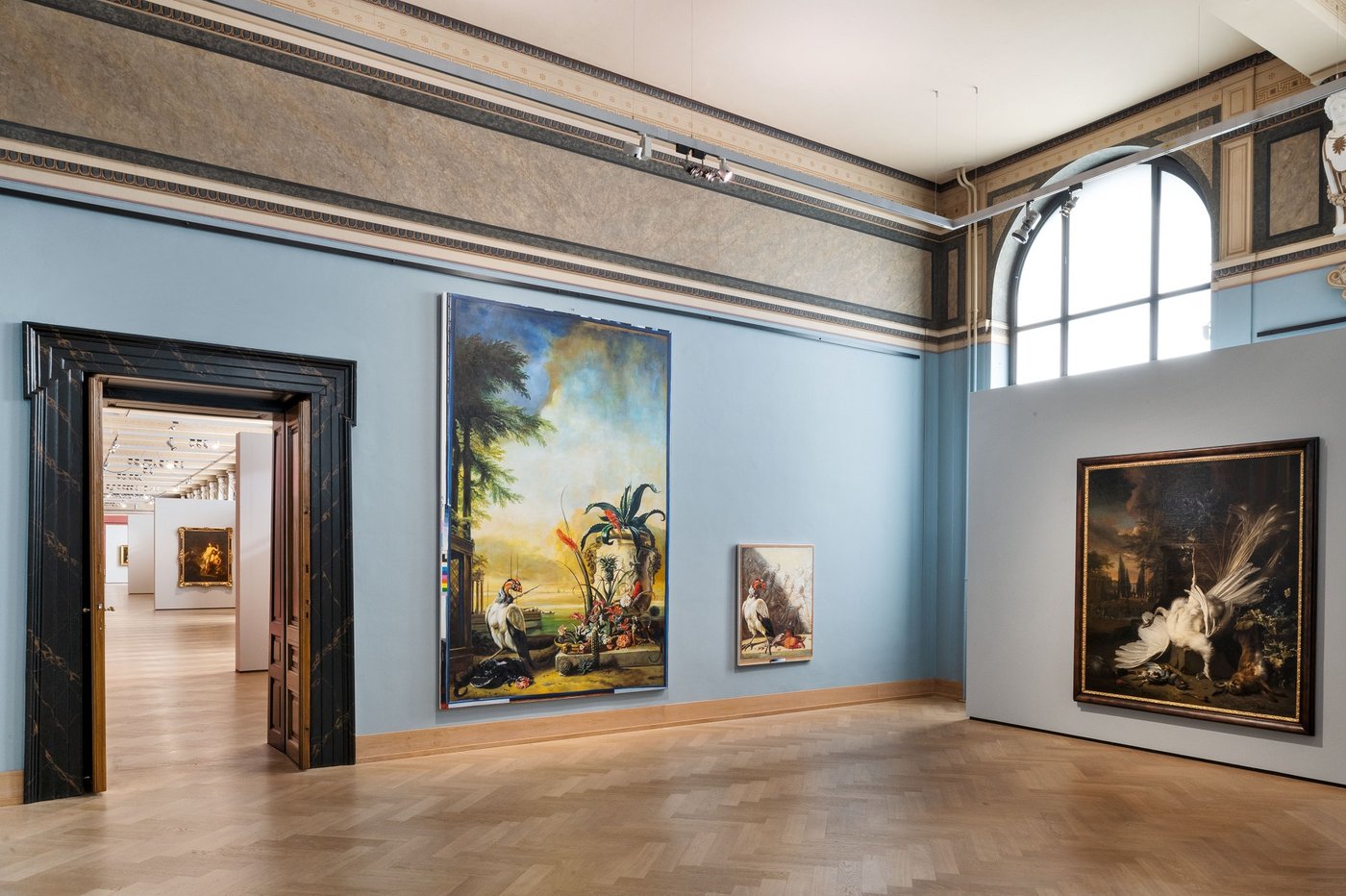 Ein blau ausgemalter historischer Ausstellungsraum mit drei bunten Gemälden und ein Blick durch eine Tür offenbar eine Einsicht in weitere weitläufige Ausstellungsräume.