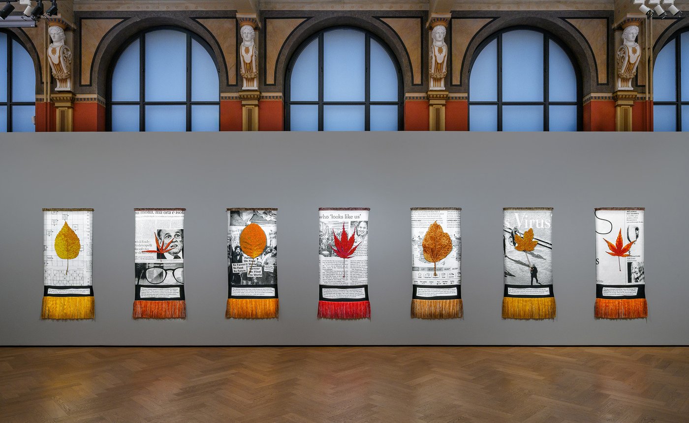 ein Austellungsraum in einem historischen Gebäude mit braunem Holzparkettboden und einer grauen Wand mit bunten Teppichen an der Wand