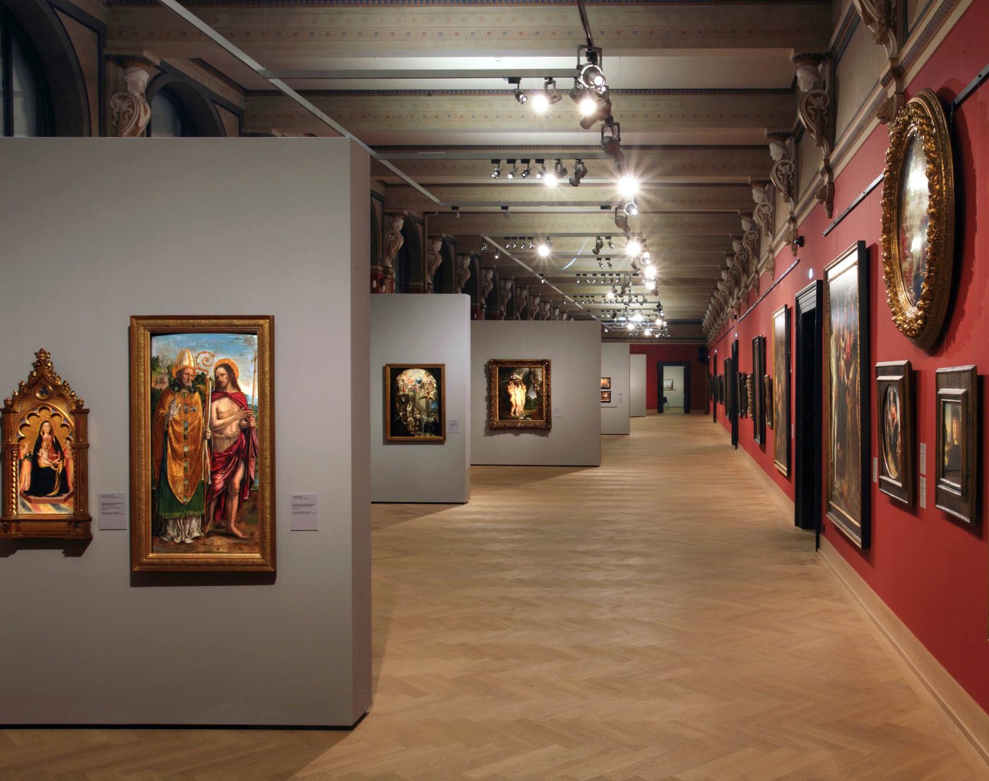 The photo shows one of the rooms of the Picture Gallery of the Academy of Fine Arts Vienna at the Schillerplatz. The room is long and tapered towards the back. There are reflectors on the ceiling which illuminate the individual paintings on the walls. On the right side is a long red wall with many pictures hanging next to and above each other. In the left half of the picture there are five grey partition walls parallel to each other, positioned at right angles to the wall on the left side. Paintings with golden frames are hanging on these walls as well. Between the parallel partitions and the right wall is a corridor leading to the door at the back.