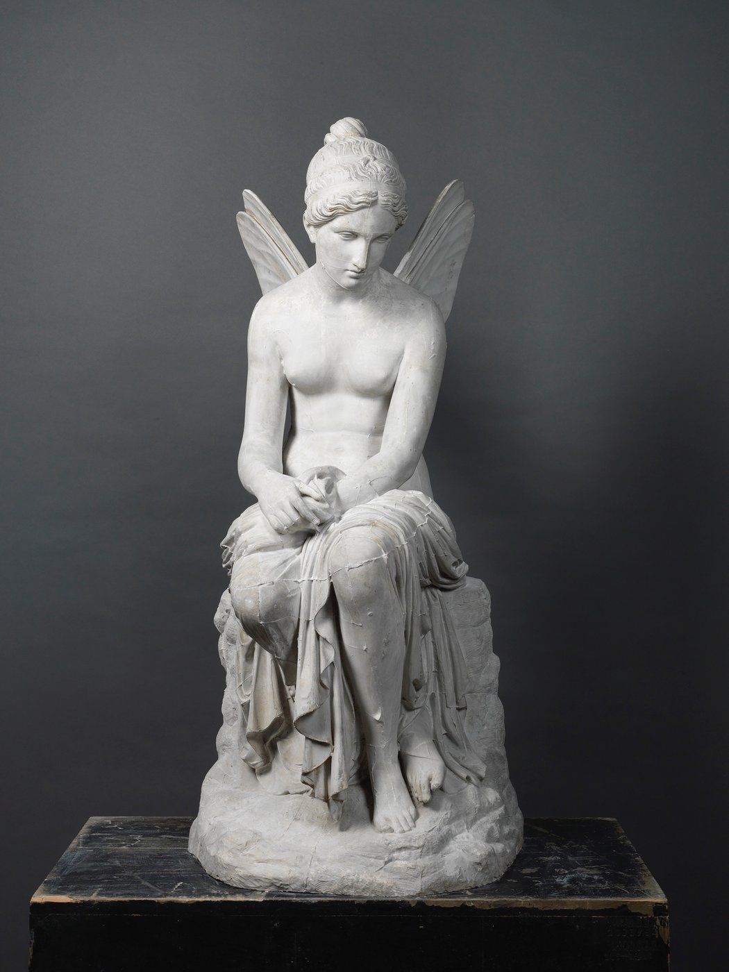 This black-and-white photograph presents a frontal view of the cast of a sculpture entitled Psyche Abandoned. The sculpture consists of a nude seated young woman with delicate wings on her back. 