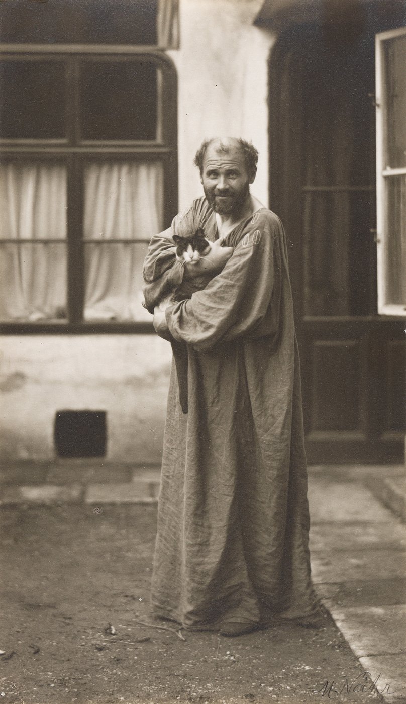 Schwarz-Weiß-Fotografie des Malers Gustav Klimt in bodenlangem Gewand und mit Katze in seinen Armen.