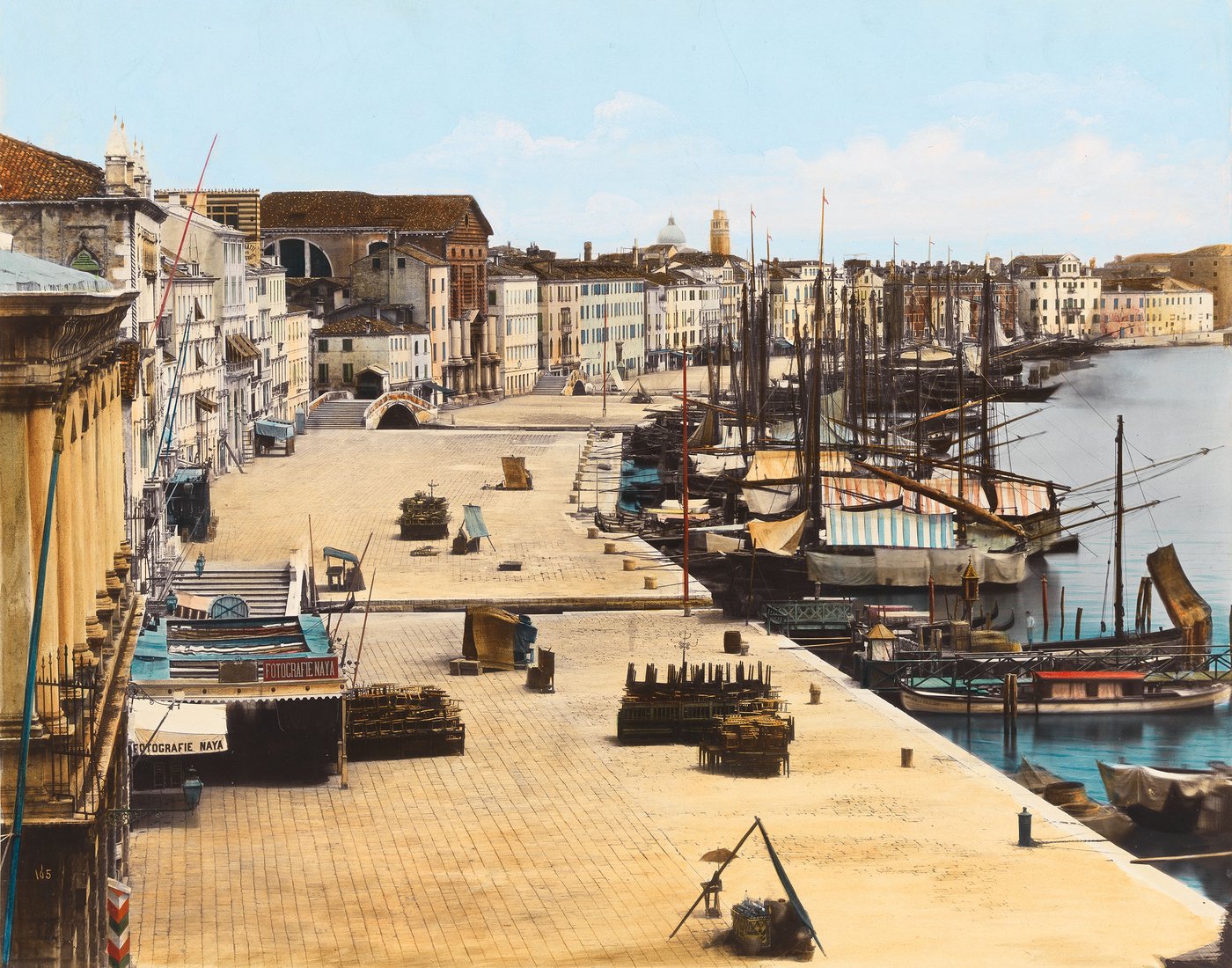 Koloriertes Foto der Uferpromenade Riva degli Schiavoni in Venedig, die von einer Häuserreihe links und der Lagune mit Segelschiffen rechts gesäumt wird. Links im Vordergrund ist das Geschäft des Fotografen zu erkennen.