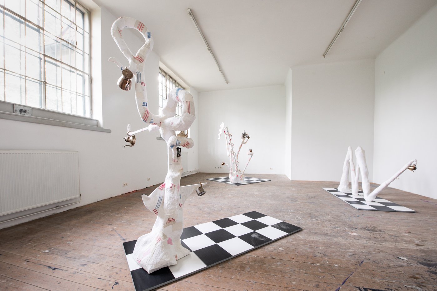 three tendril-like white plaster figures in which teapots hang, on black and white checkered surfaces