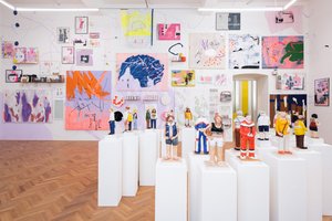 Photo of an exhibition space with small colorful figures on white pedestals, in the background colorful drawings and paintings on the wall.