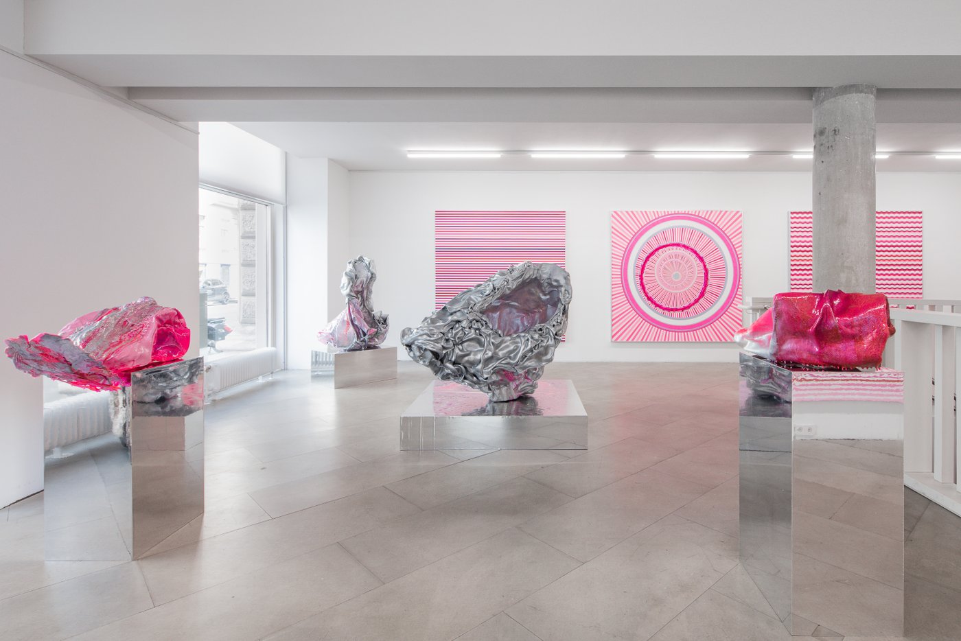 4 silver metallic objects that look like crumpled aluminum foil on silver pedestals, two of them are dipped in pink color, in the background on the wall three large-sized paintings with pink-white lines and circles