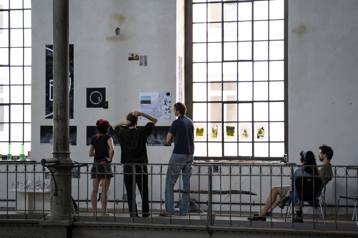 mehrere Personen auf einer Galerie mit Geländer und großen hellen Fenstern schauen Kunstwerke an der Wand an, rechts im Bild sitzen zwei Personen