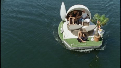 eine Schwimminsel im Wasser mit einer Palme, Bänken mit Sonnenschutz auf dem Personen sitzen