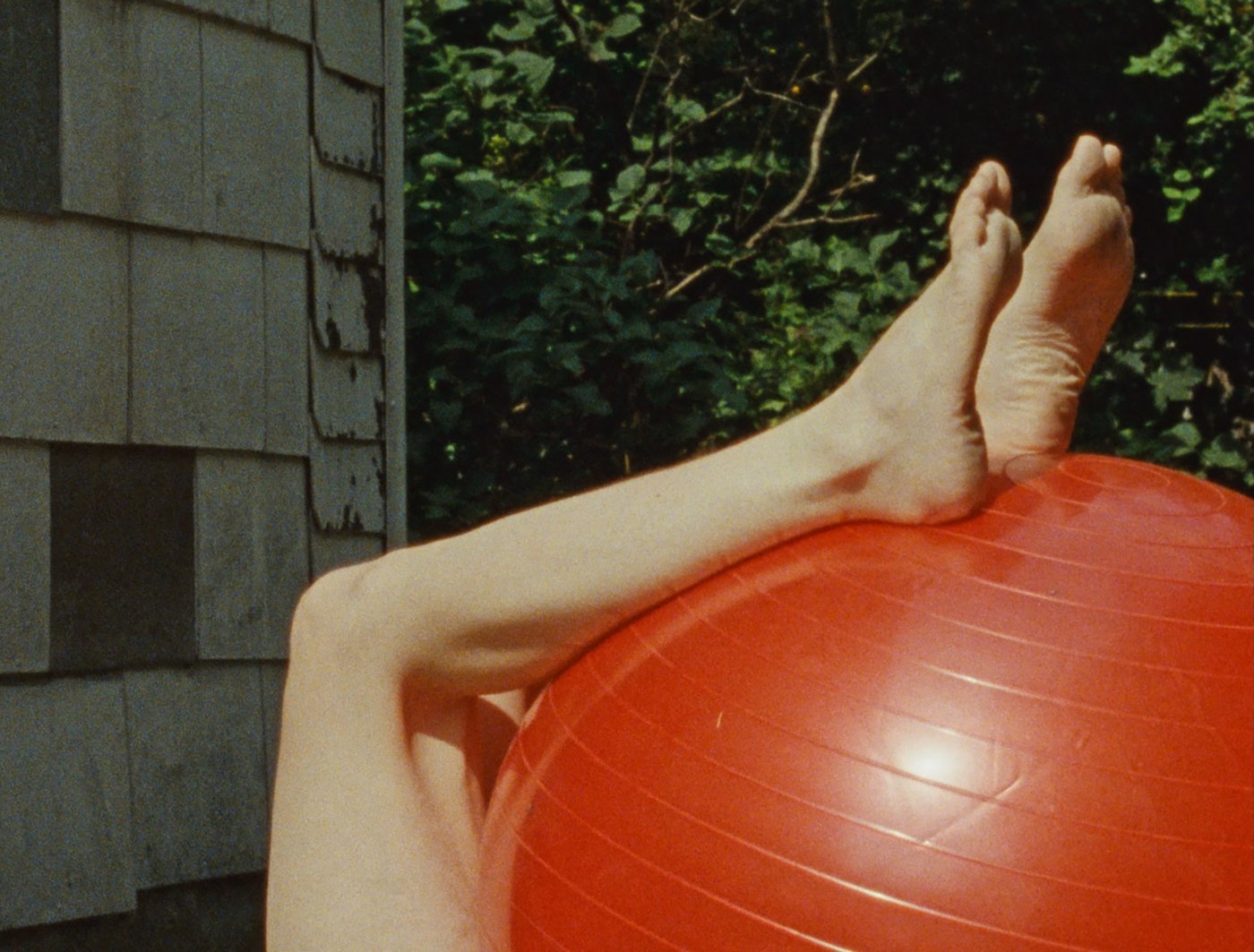 Fotografie von Beinen die auf einem roten Gymnastikball lehnen, im Hintergrund eine grüne Hecke und ein Teil einer Hauswand