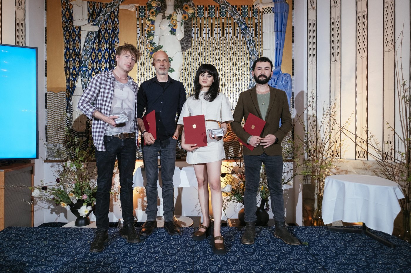 Foto der vier Preisträger_innen, zwei Männer, eine Frau in der Mitte und ein Mann rechts von ihr, alle halten eine rote Urkunde in ihren Händen, sie stehen nebeneinander vor einem Klimt Wandbild auf einem dunkelblauem Teppichboden