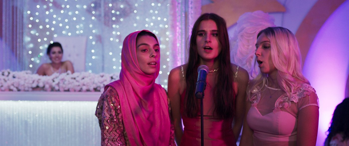 Filmstill des Filmes "Sonne" auf dem drei junge Frauen zu sehen sind die zusammen in ein Mikrofon singen, sie sind festlich gekleidet, das Bild ist in ein Rosa Licht getaucht