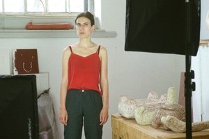 Foto einer Frau in rotem Top mit dunkler Hose, steht in einem Raum vor einem Fenster mit verschiedenen Objekten um sie herum