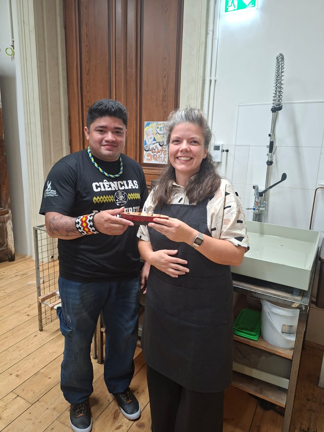 Zwei Personen stehen in einer Werkstatt, halten ein Werkstück und lachen in die Kamera.