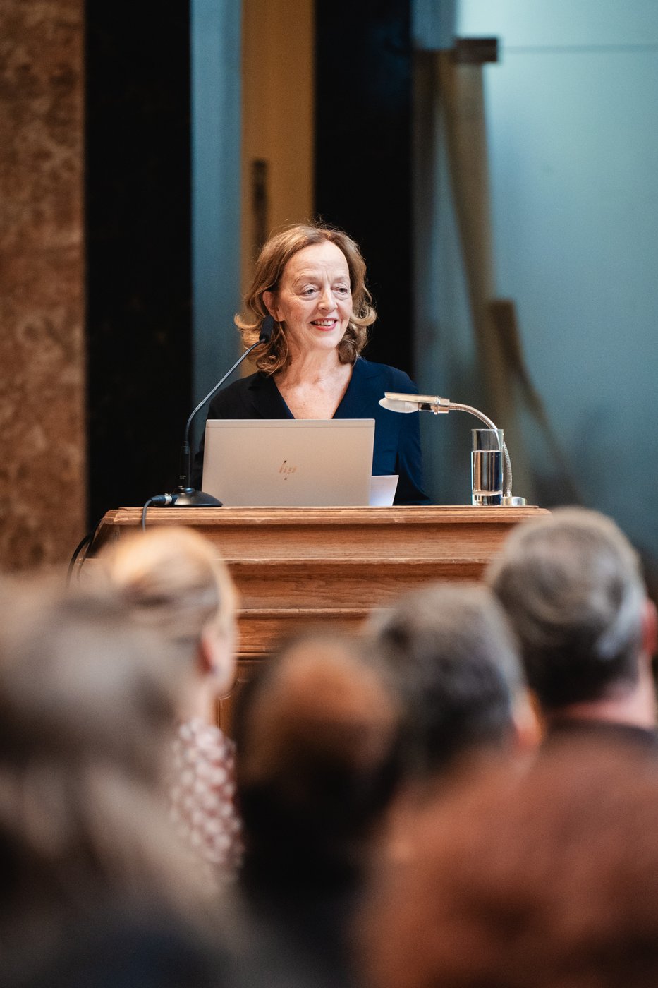 Person steht hinter einem Pult vor Publikum und hält einen Vortrag.