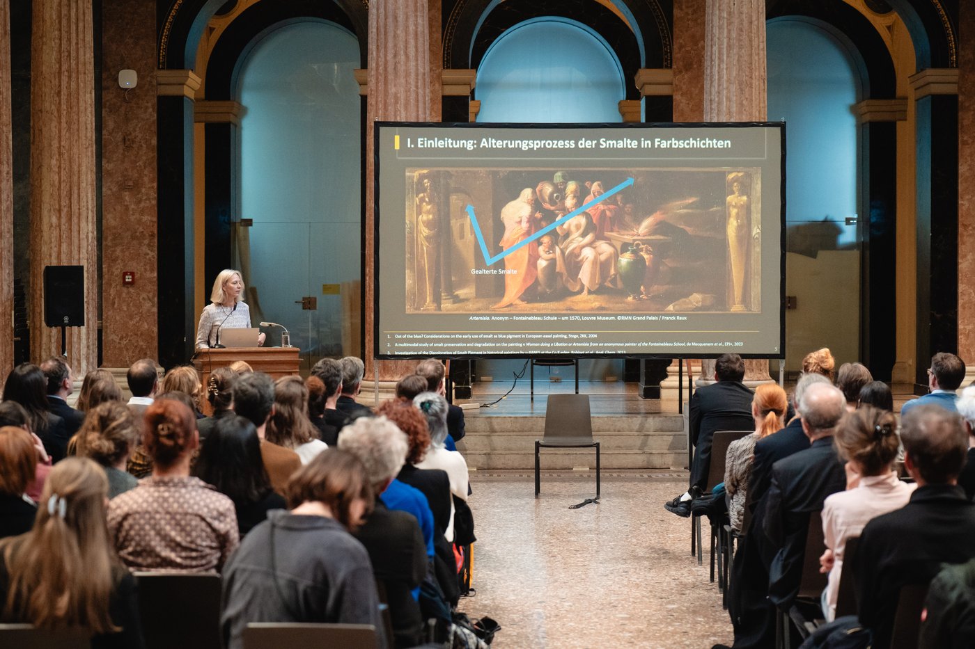Eine Person steht an einem Pult und hält einen Vortrag, neben ihr auf einer Leinwand eine Power-Point-Folie. Vor ihr sitz das Publikum.