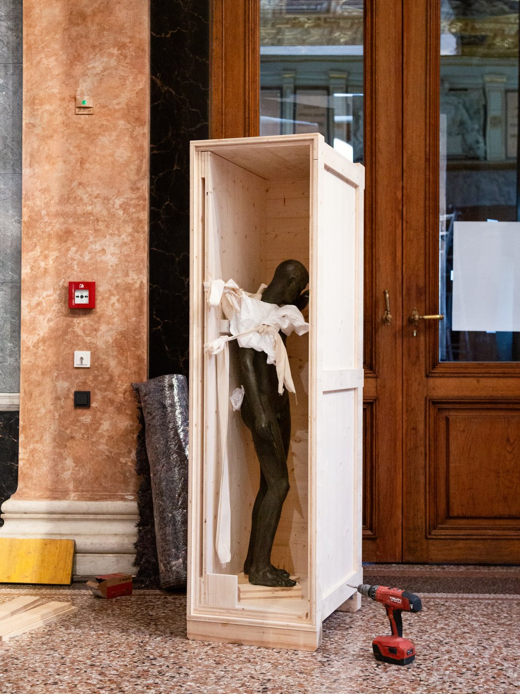 Eine schwarze Statue ist in einer hohen, zu einer Seite offenen Box aus Holz zu sehen.Daneben stehen eine Bohrmaschine und Material für den Kunsttransport.