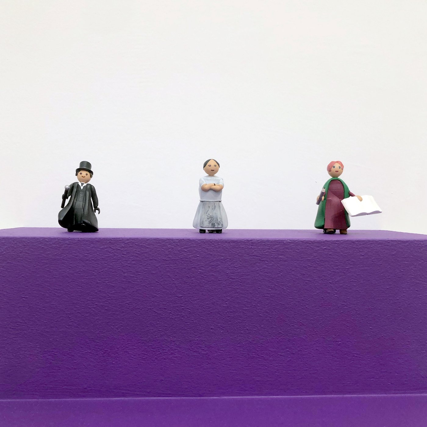 drei kleine Holzfiguren auf einem Lilafarbigen Podest, links ein Mann in schwarzem Anzug mit Stock und Zylinder, in der Mitte ein Frau in weißem Kleid mit verschänkten Armen, rechts eine Frau mit weinrotem Kleid, gründem Schal und einem Buch in der Hand