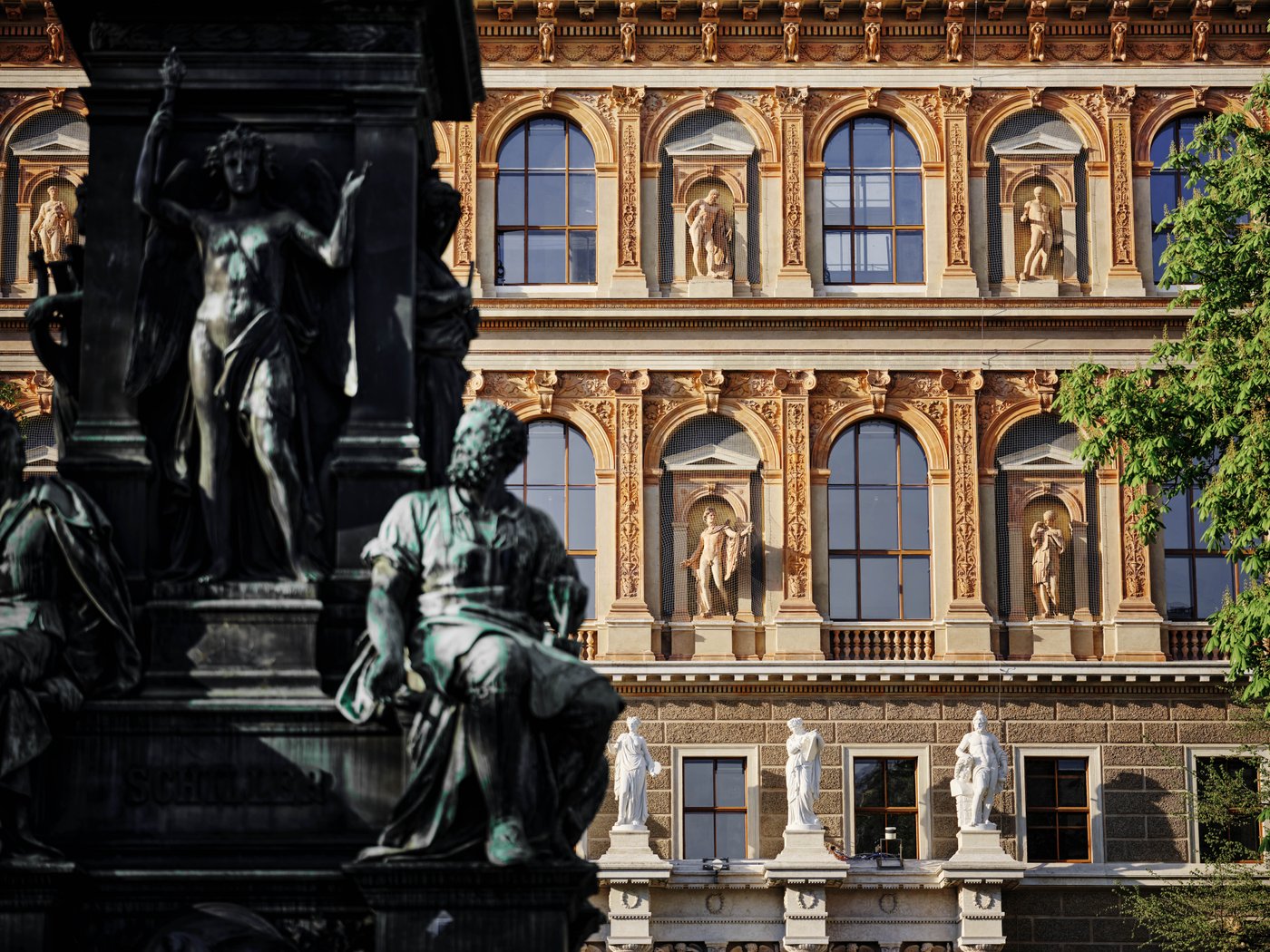 Nahaufnahme eines hisotirschen Gebäudes, Ansicht der Fassade mit Bogenfenster und Statuen in Giebeln, im Vordergrund unscharf Detail einer Bronzestatue