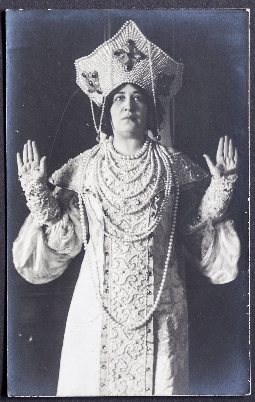 schwarz-weiß Foto einer Frau in historischem Gewand mit kronenförmiger Kopfbedeckung, die Arme mit flachen Händen abgewinkelt gehoben