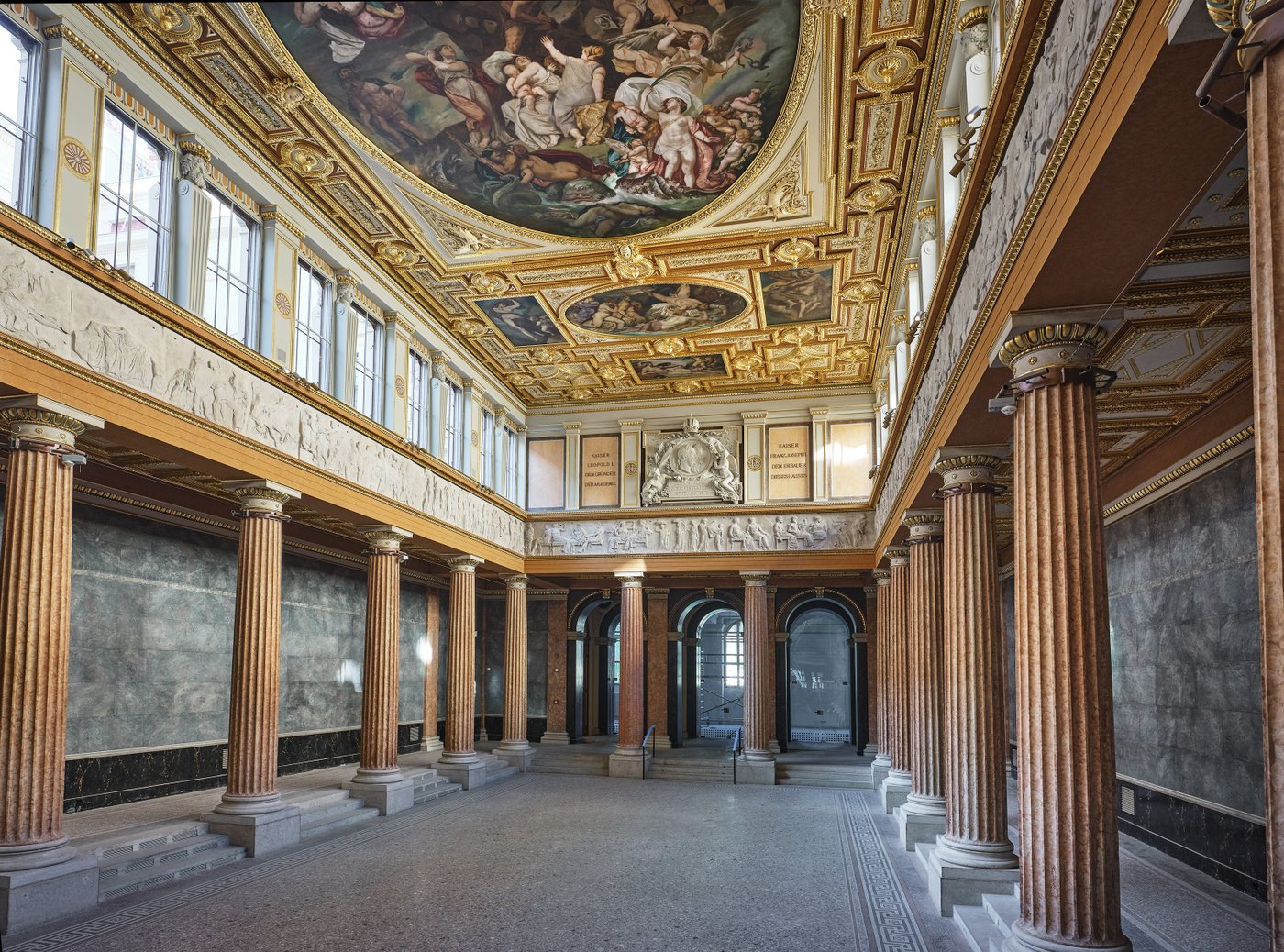 Ansicht einer Aula mit roten griechischen Säulenreihe, Stufen, figuralem Fries,goldverzierten eckigen Oberfenstern, gold verzierte Decke mit Stuckdeko die Deckenmalerei umfasst
