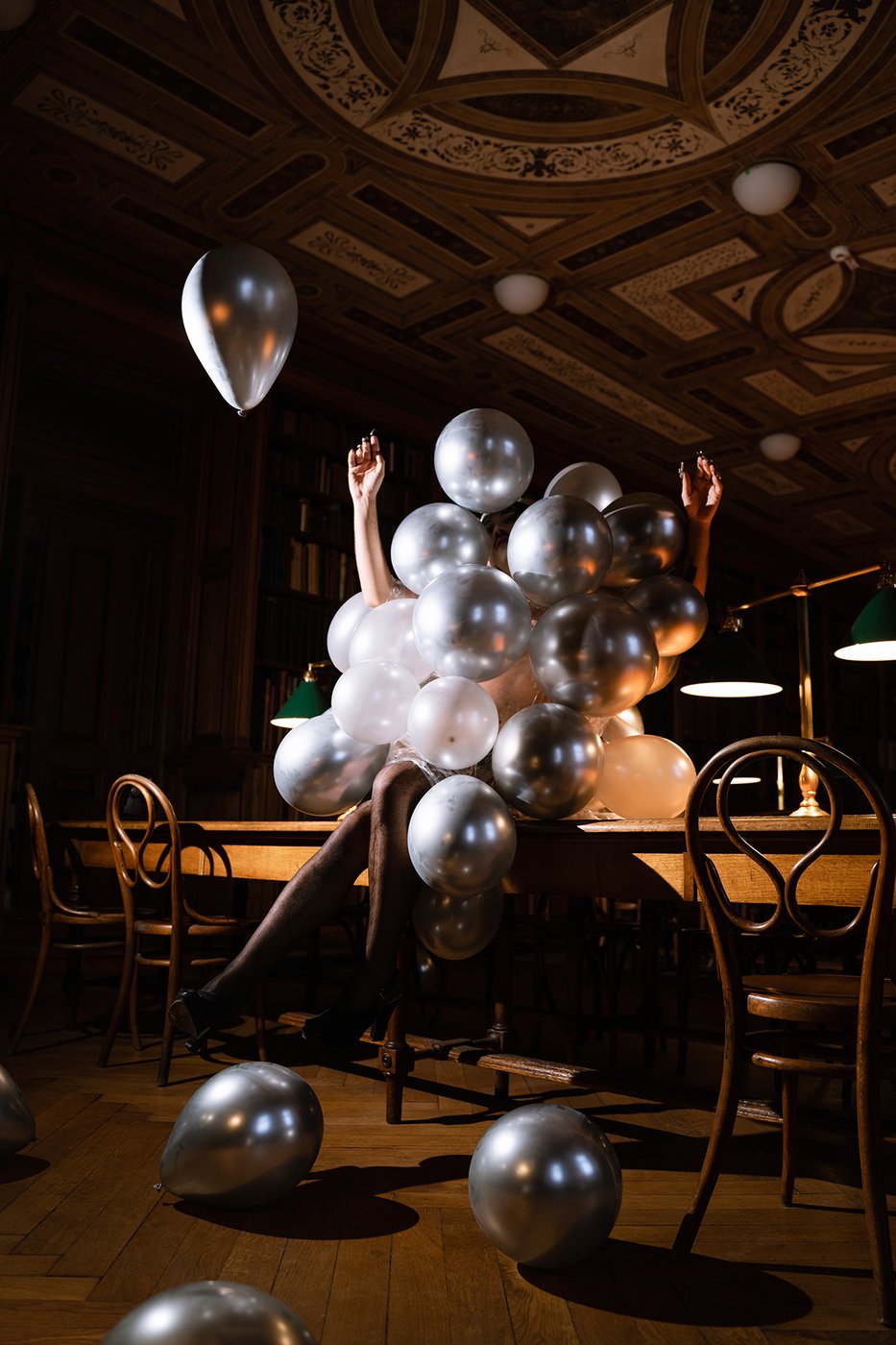 dunkler Raum mit holzverkleidung, auf einem Holztisch sitz eine Person die von silbernen Luftballonen umhüllt ist, diese fliegen auch im Raum herum
