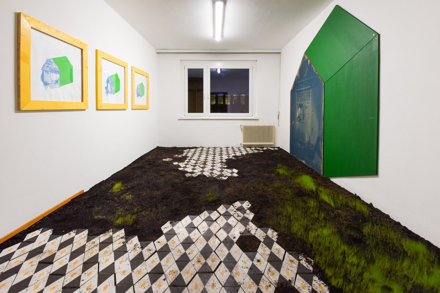 In einem Raum wächst Gras aus dem Boden der teilweise mit Fliesen ausgelegt ist, an den Wänden hängen bunte Bilder
