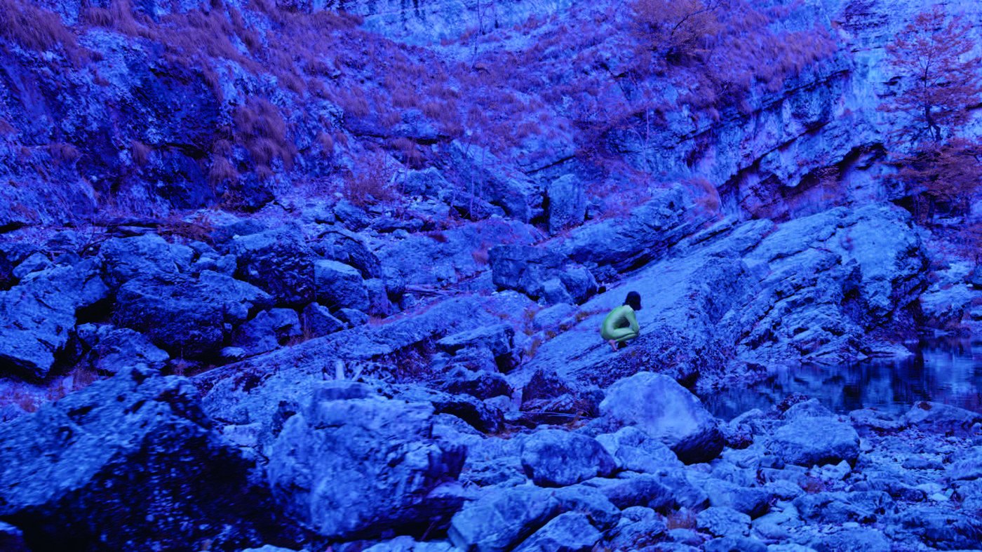 Eine nackte Person hockt in einer wüstenartigen Landschaft