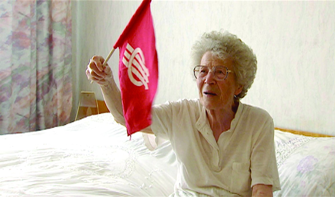 Eine alte Frau sitzt auf einem Bett und schwingt eine rote Fahne mit dem Zeichen der Sozialdemokratie