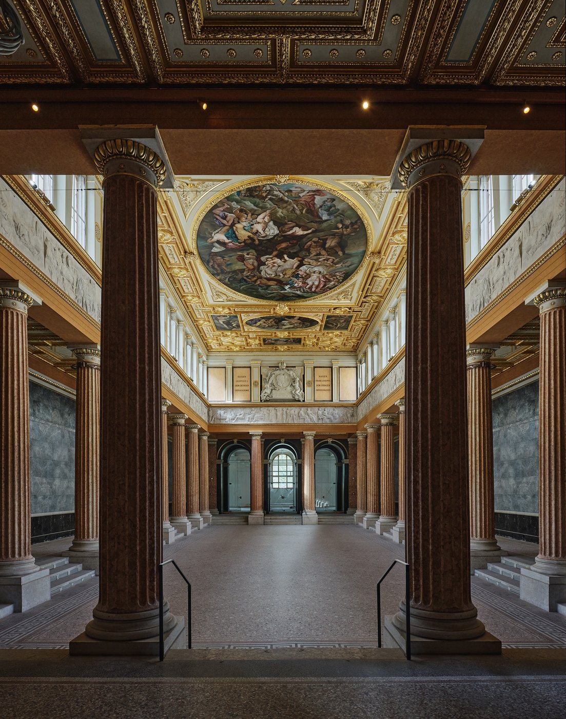 Aula mit roten Säulenarkarden und goldener verzierter Decke mit ovalen Deckengemälden, darunter ein Figurenfries