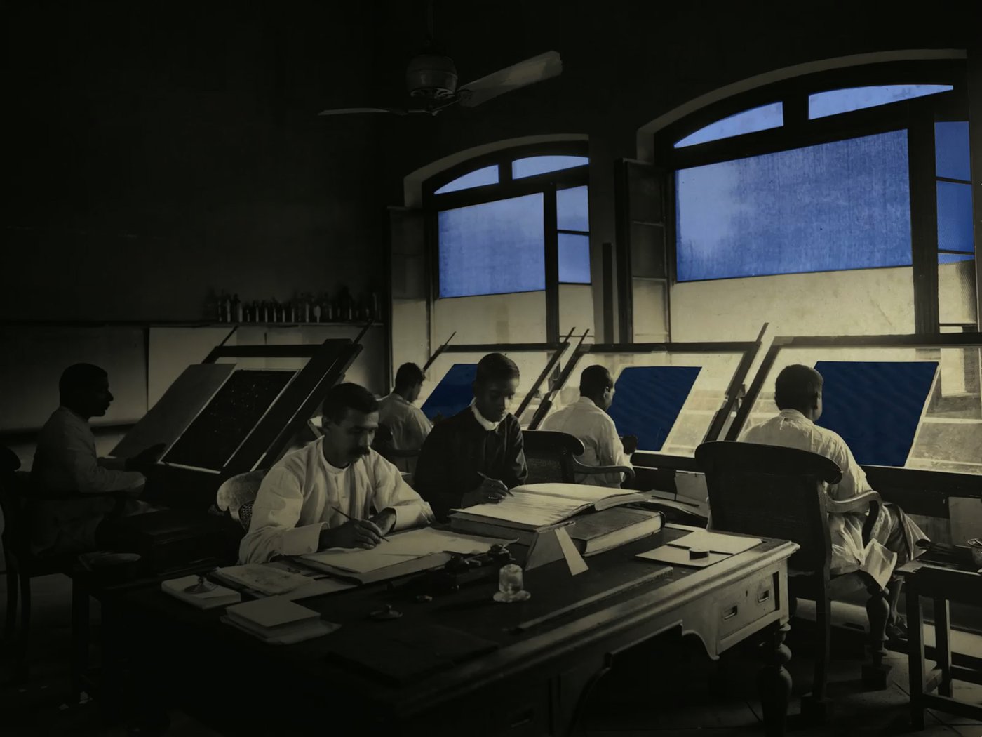 schwarz-weiß Foto von Personen in einem Arbeitsraum vor Zeichenpulten die mit blauen Akzenten hervorgehoben werden