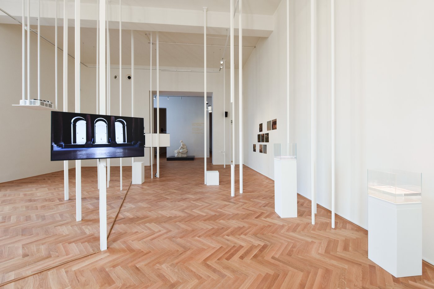 Ausstellungsansicht mit weißen senkrechten Stäben durch den Raum und einem Bildschirm im Vordergrund, an der Wand im Hintergrund Fotos
