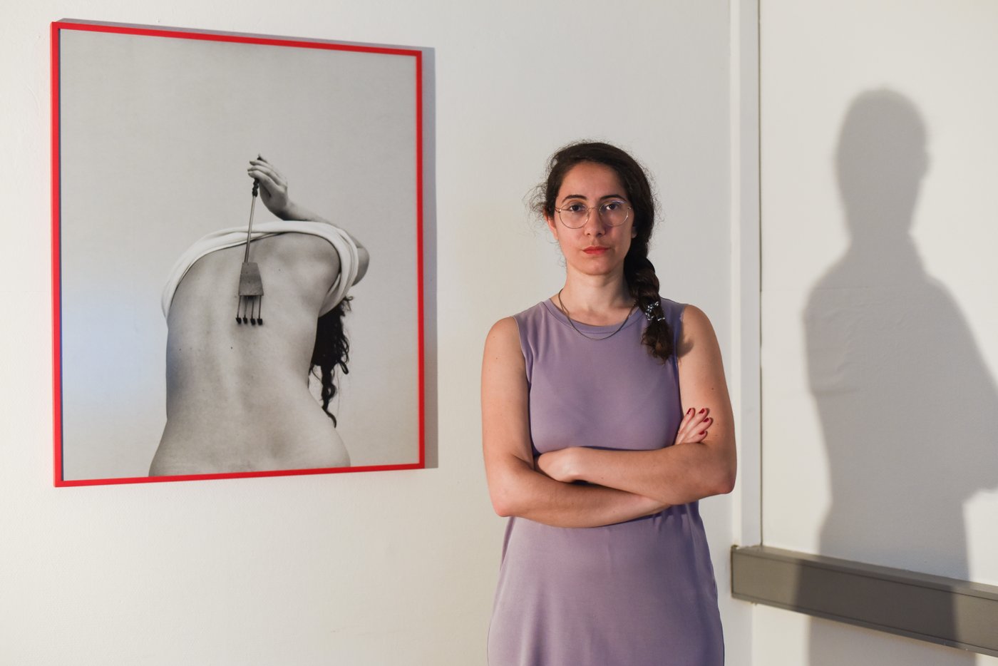 Eine Person mit dunklen Haaren in einem fliederfarbenen Kleid steht in einem Raum vor einem Foto auf dem sich eine Person mit einer Gabel den Rücken kratzt.