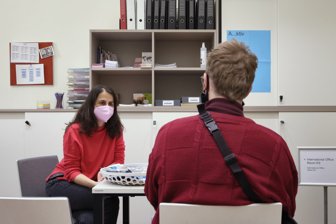 Eine Frau und ein Student sitzen an einem Tisch, beide haben rote Pullover und Masken an. Im Hintergrund sieht man typische Büroeinrichtung.
