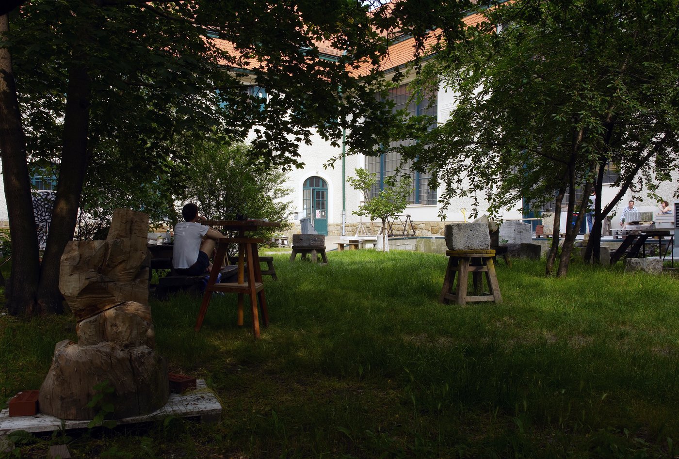 In einem sommerlichen Garten sitzt ein junger Mann, den Rücken zugewandt, die Beine angezogen, mit einem weißen T-Shirt. Er ist umgeben von Holz- und Steinskulpturen. Im Hintergrund sieht man das historische Gebäude mit den großen Atelierfenstern.