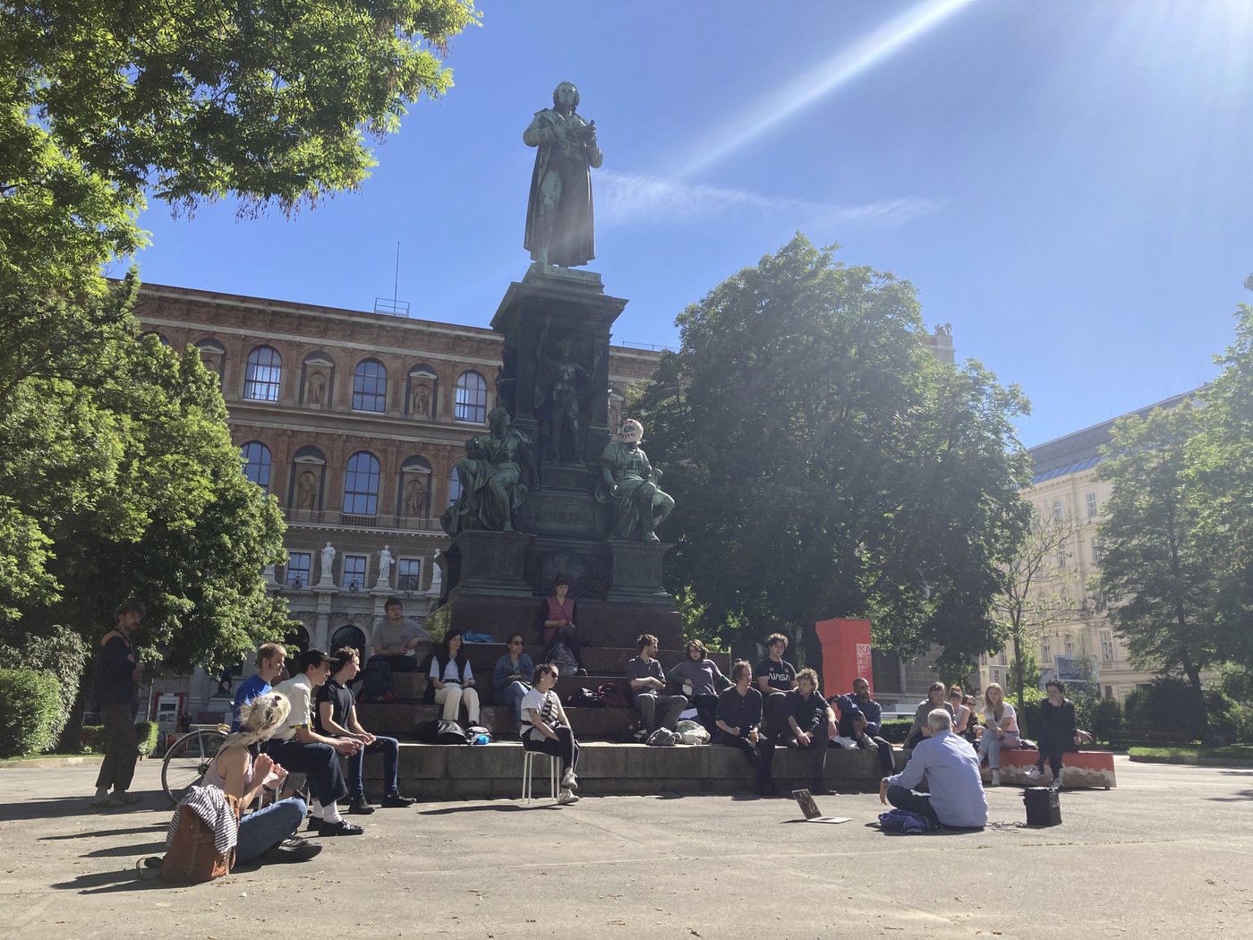 Das Bild zeigt eine Gruppe von Studierenden, die auf dem Sockel des Schillerdenkmales sitzen, offensichtlich in einer Unterrichtsituation. Es ist Sommer und man sieht das barocke Denkmal mit Friedrich Schiller, einige Bäume und dahinter das Gründerzeitgebäude der Akademie.