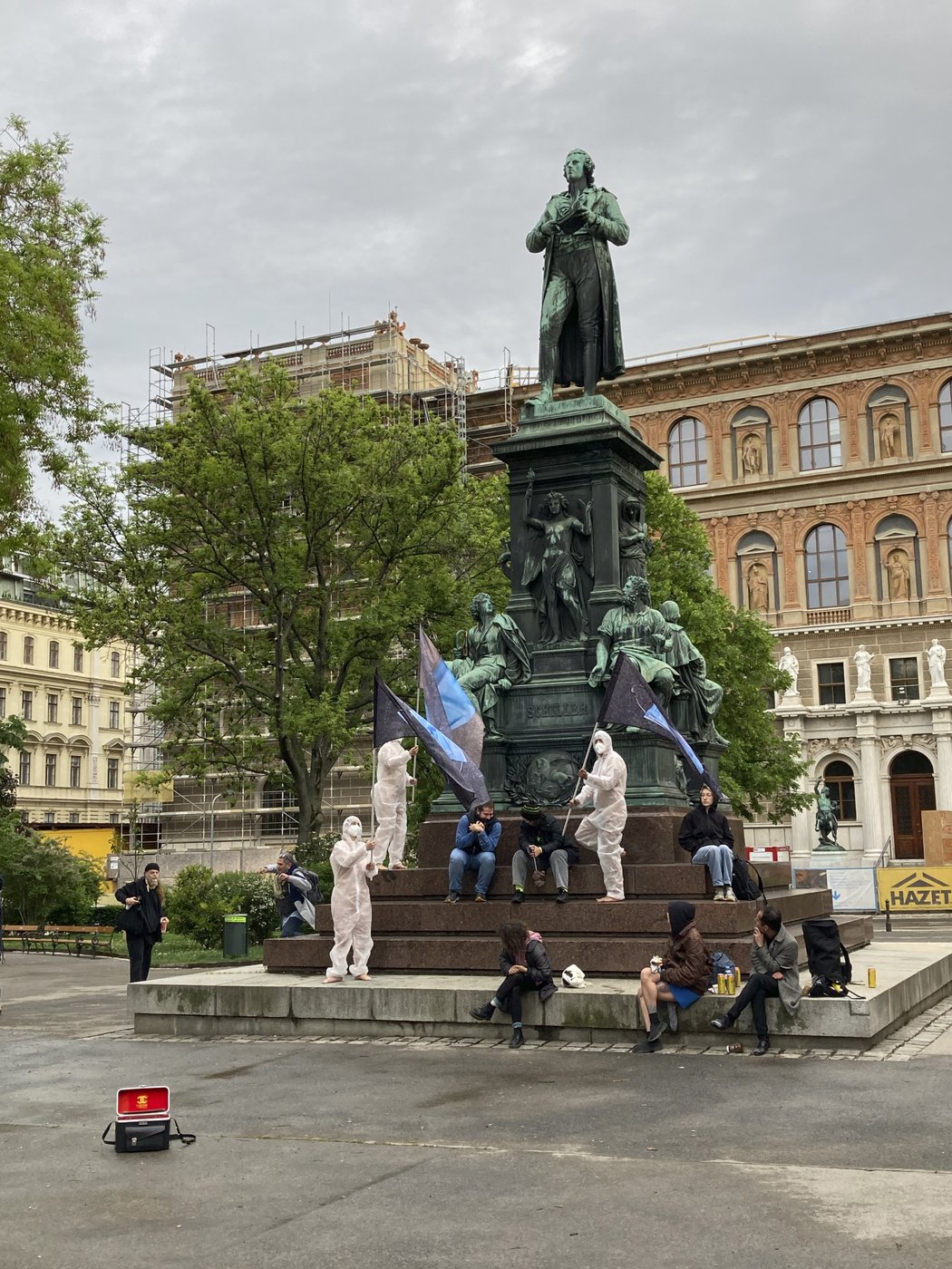 Das Bild zeigt eine Gruppe von Studierenden, die auf dem Sockel des Schillerdenkmales sitzen. Drei von ihnen haben weiße Anzüge und FFP2 Masken an, sie tragen schwarze Fahnen mit einem blauen Zeichen.