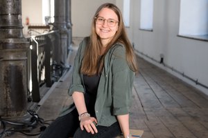 Person langen Haaren und Brille trägt eine grüne Bluse und sitzt auf einem Hocker und lächelt in die Kamera