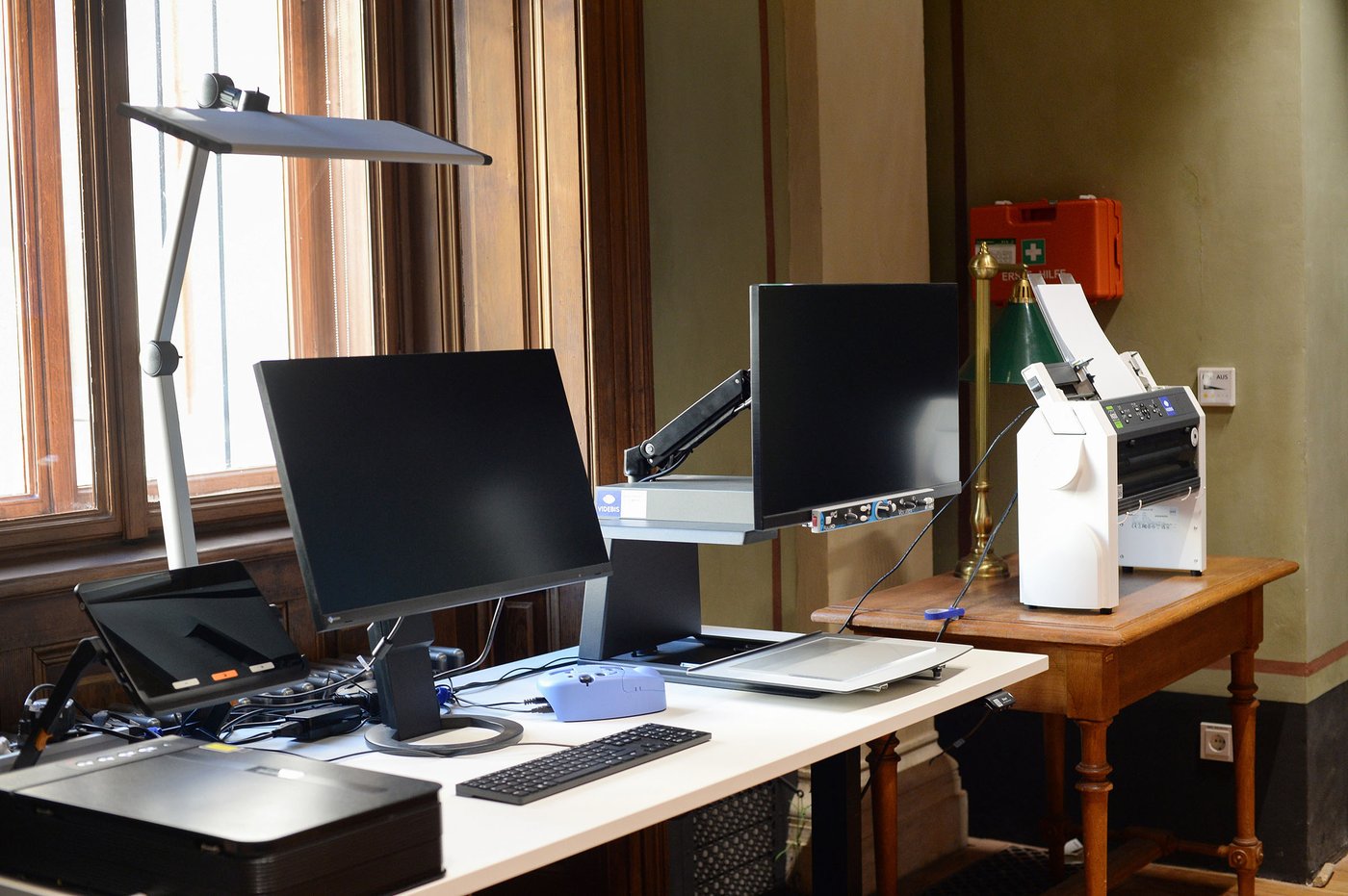 Die Farbfotografie im Querformat zeigt den barrierefreien Arbeitsplatz im Seitengang der Universitätsbibliothek u. a. mit folgenden Geräten: Monitor, Scanner, Bildschirmlesegerät, Brailledrucker.